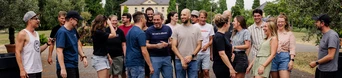A diverse group of people gathered outdoors, engaging in conversation and laughter, with a building and trees in the background.