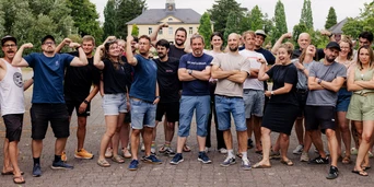 Group of diverse individuals posing together outdoors, with some flexing muscles and others crossing arms, in front of a building and trees.