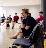 A woman sits on a chair, smiling and engaged, while a group of people are seated in a circle in a well-lit room.